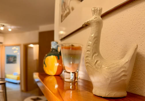 Ceramic figurine of a bird on a wooden shelf, with a colorful glass and a small yellow vase in the background, in a warmly lit room.