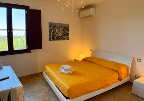 A bedroom with a double bed covered in a yellow blanket, two bedside tables with lamps, a wall-mounted air conditioner, a window with open shutters, a painting on the wall, and a dresser with a TV on top.