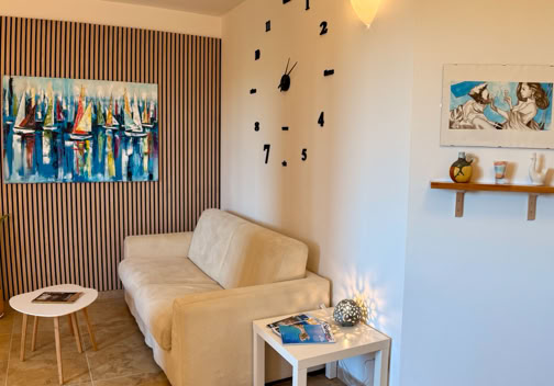 Salotto con divano beige, tavolino bianco, pianta in vaso, orologio da parete nero, quadro astratto e mensola con decorazioni.