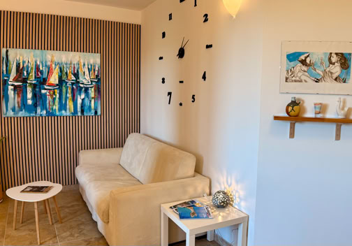 A cozy living room with a beige sofa, a small round white table, and a side table with a book. A large abstract painting and a wall clock are on the wall. A small shelf holds decorative items, and a potted plant is in the corner.