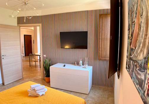 A modern bedroom with a yellow bedspread, a wall-mounted TV, a white cabinet, and a painting on the wall. A door is open, revealing a hallway. A plant and folded towels are on the cabinet.
