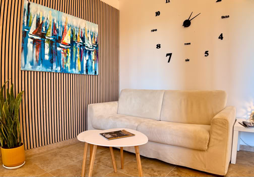 A living room with a beige sofa, a small white coffee table, a wall clock with black numbers, a colorful abstract painting, a potted plant, and a side table with a decorative item.