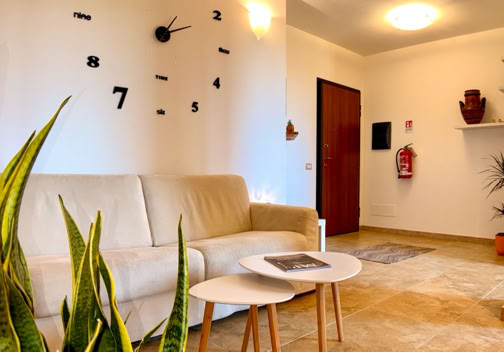 Soggiorno con divano beige, due tavolini bianchi, pianta verde, orologio da parete nero, porta d'ingresso e decorazioni su mensola.